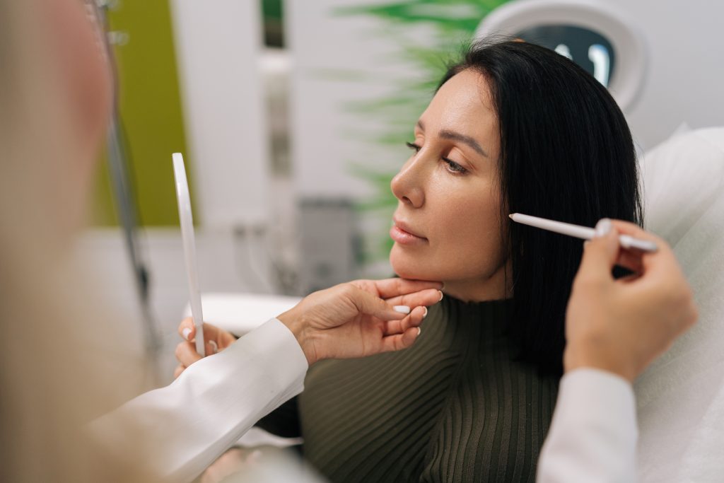 Side view of woman looking in mirror while cosmetologist marking face for aesthetic procedure, preparing for beauty treatment in cosmetology clinic. Concept of facial anti aging, skincare treatment – Alpha Hospital Group